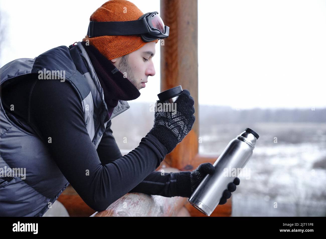 Un ragazzo in un cappello arancione in inverno beve il tè da un thermos durante una vacanza invernale in montagna. Si trova sul balcone di una casa di legno. Porta Foto Stock