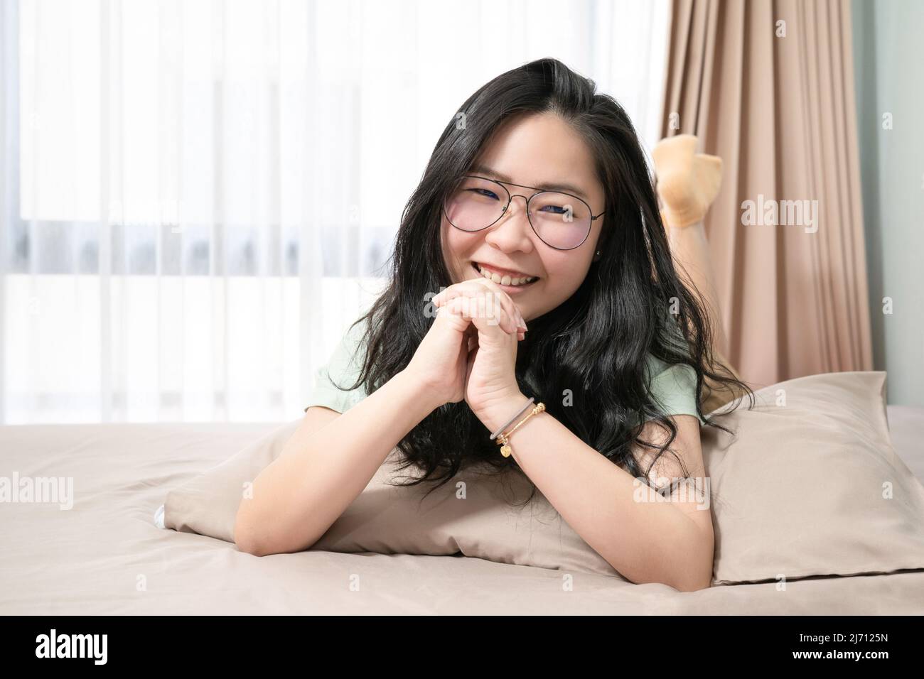 Bella donna asiatica tenere le mani insieme mentre si sdraia sul suo stomaco sul letto in camera da letto con un tema di colore verde pastello - marrone. Foto Stock