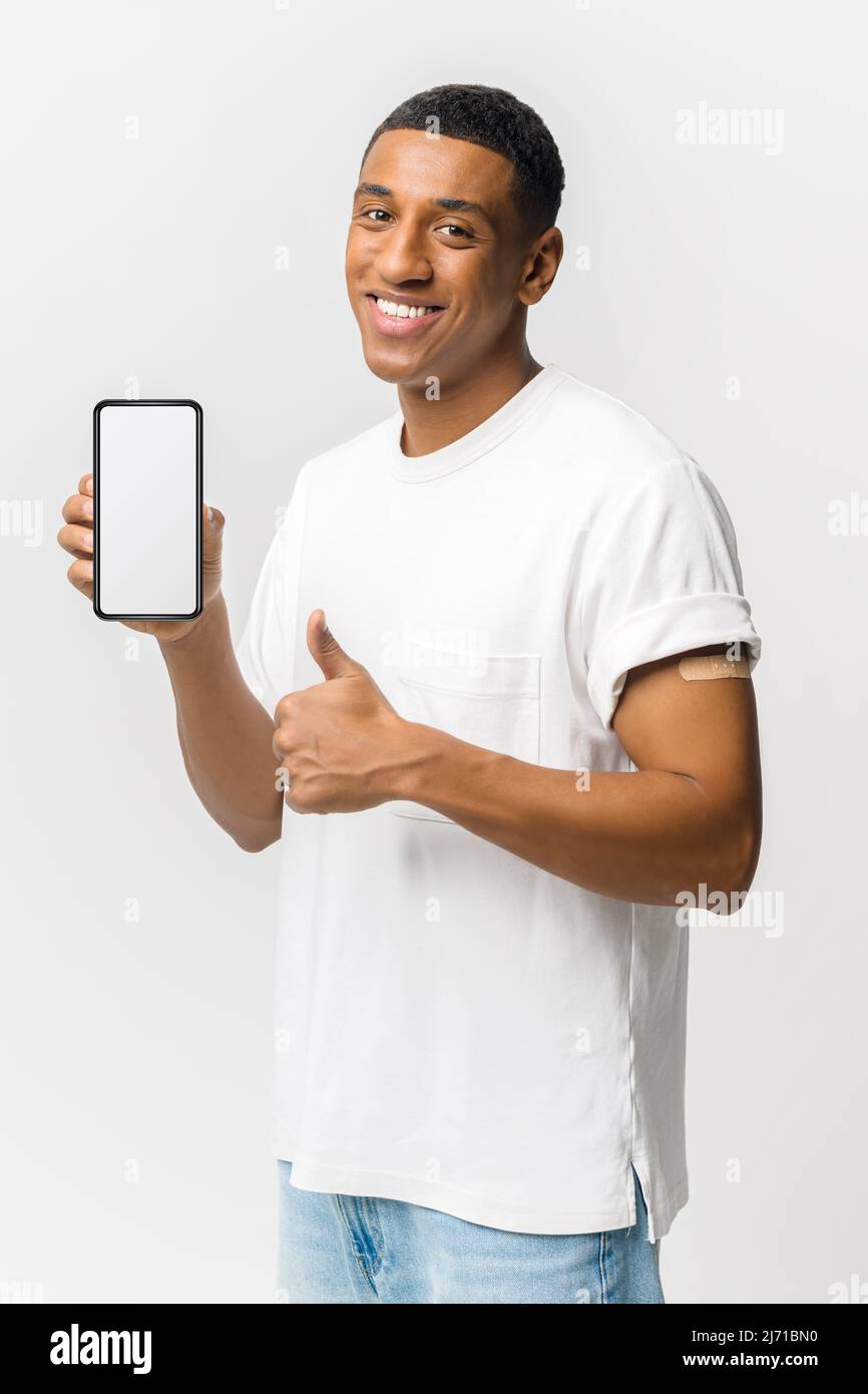Sorridendo giovane ragazzo latino con band-aid dopo l'iniezione del vaccino sul braccio mostrando smartphone con schermo vuoto e pollice su isolato su sfondo blu, mock-up concetto Foto Stock