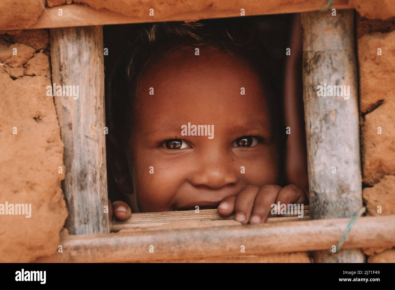 Bambino colpito dalla povertà che guarda e sorride attraverso la piccola finestra di una casa di fango nel Nord del Brasile. Foto Stock