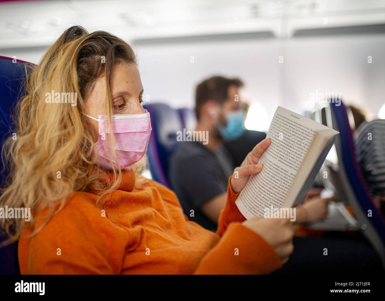 Donna matura che legge in aereo di tipo pandemico con maschera rosa che indossa l'arancione e i passeggeri che indossa le maschere sullo sfondo Foto Stock