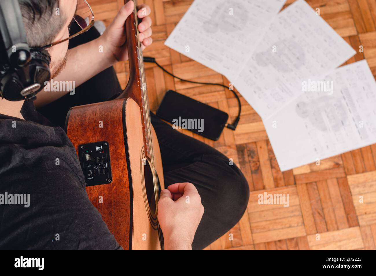 vista dall'alto, uomo seduto sul pavimento che studia musica con chitarra acustica, cuffie, spartiti musicali e un telefono sul pavimento. Foto Stock