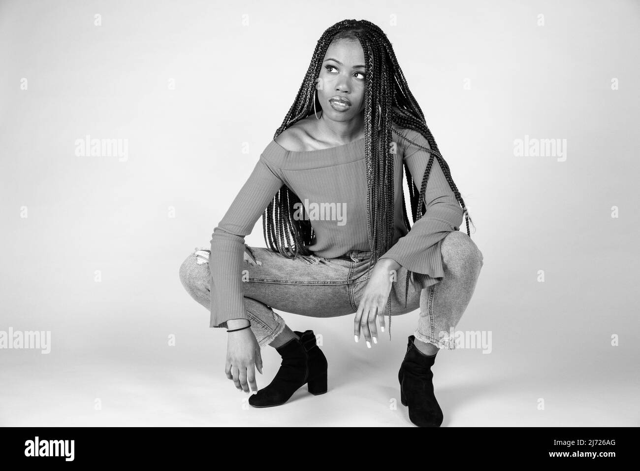 Studio shot di una giovane donna nera con vintage top arancione e i jeans larghi in posa su uno sfondo bianco squatting Foto Stock