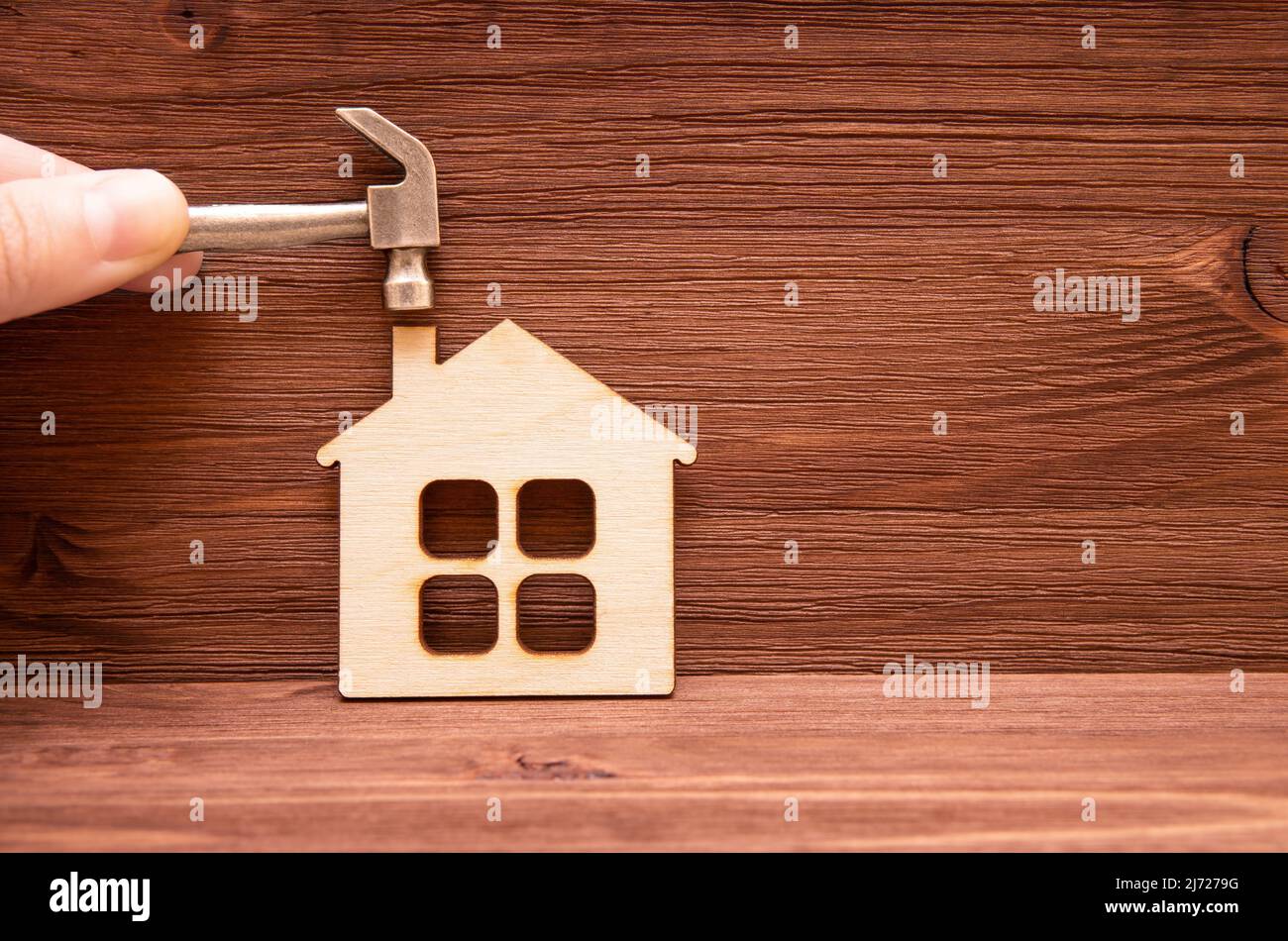 Dita che tengono un martello in acciaio miniaturizzato su un piccolo modello di casa piatta su uno sfondo di legno. Home progetto di revamping. Foto Stock