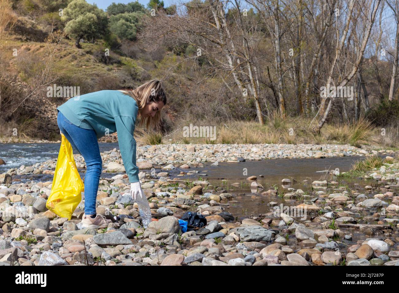 Donna accovacciata giù per prendere una bottiglia di plastica trovata fra le pietre di fiume. Concetto di ecologia Foto Stock