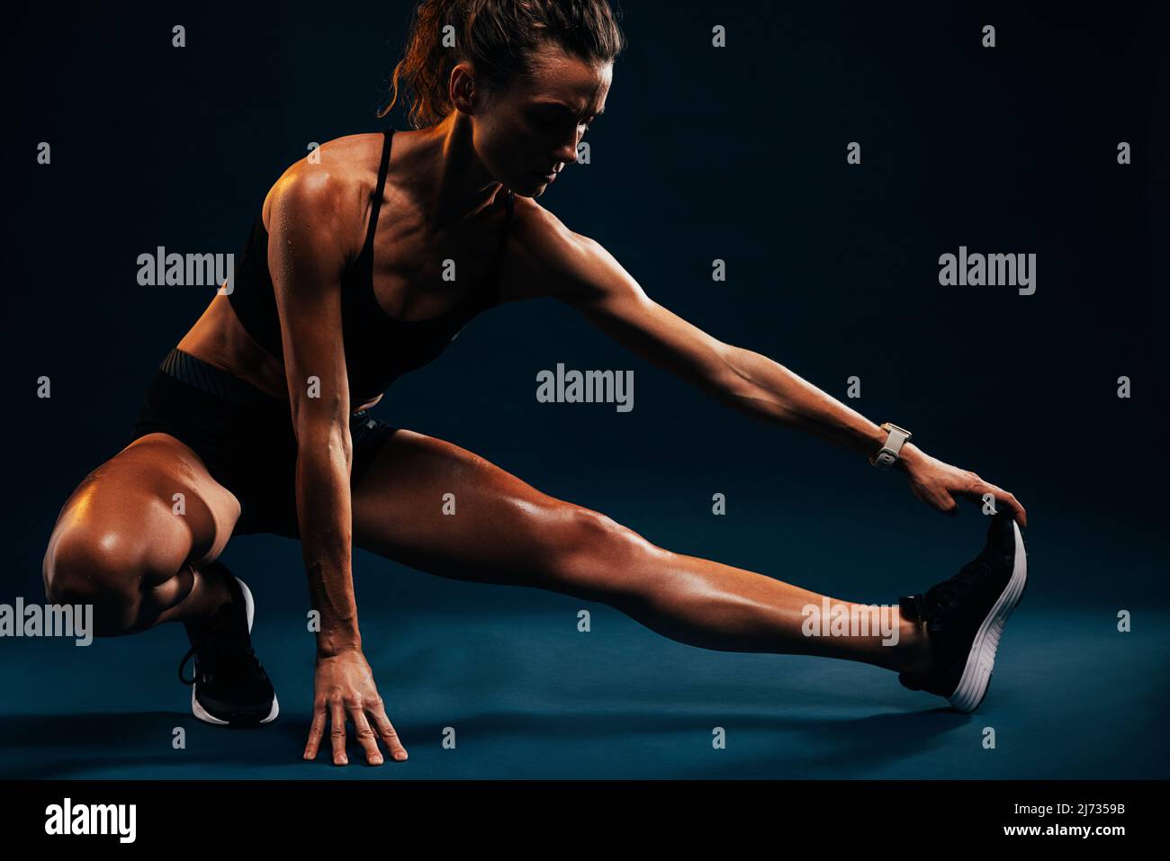Atleta che fa esercizi di stretching su sfondo nero. Donna runner che allunga i muscoli delle gambe toccando le sue scarpe mentre si siede in studio. Foto Stock