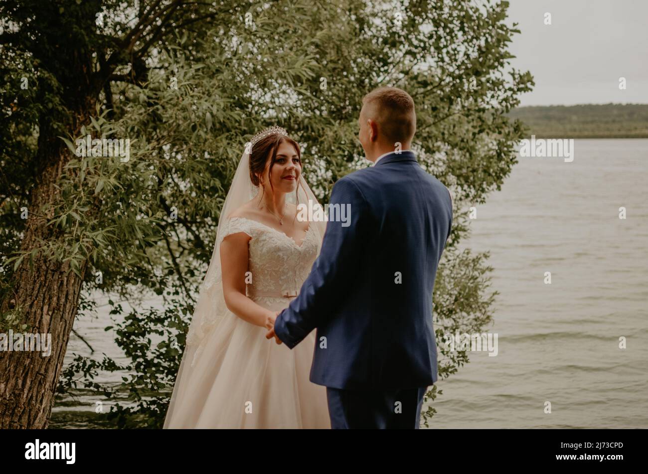 Il giovane uomo biondo caucasico europeo sposa in abito blu e donna dai capelli neri sposa in abito da sposa bianco con velo lungo e tiara sulla testa. Sposi novelli Foto Stock