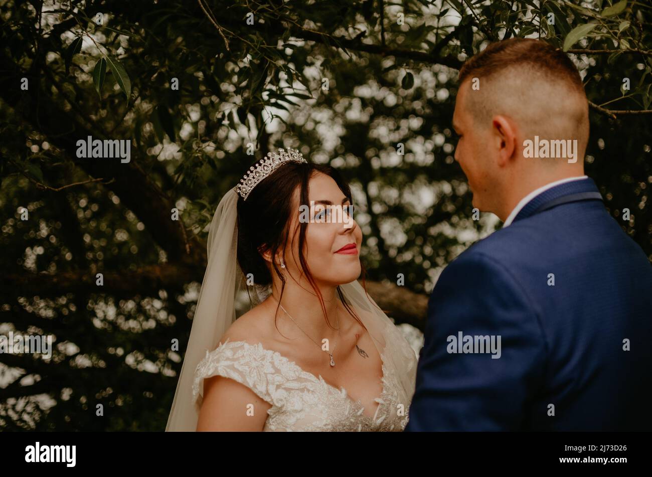 Il giovane uomo biondo caucasico europeo sposa in abito blu e donna dai capelli neri sposa in abito da sposa bianco con velo lungo e tiara sulla testa degli sposi novelli Foto Stock