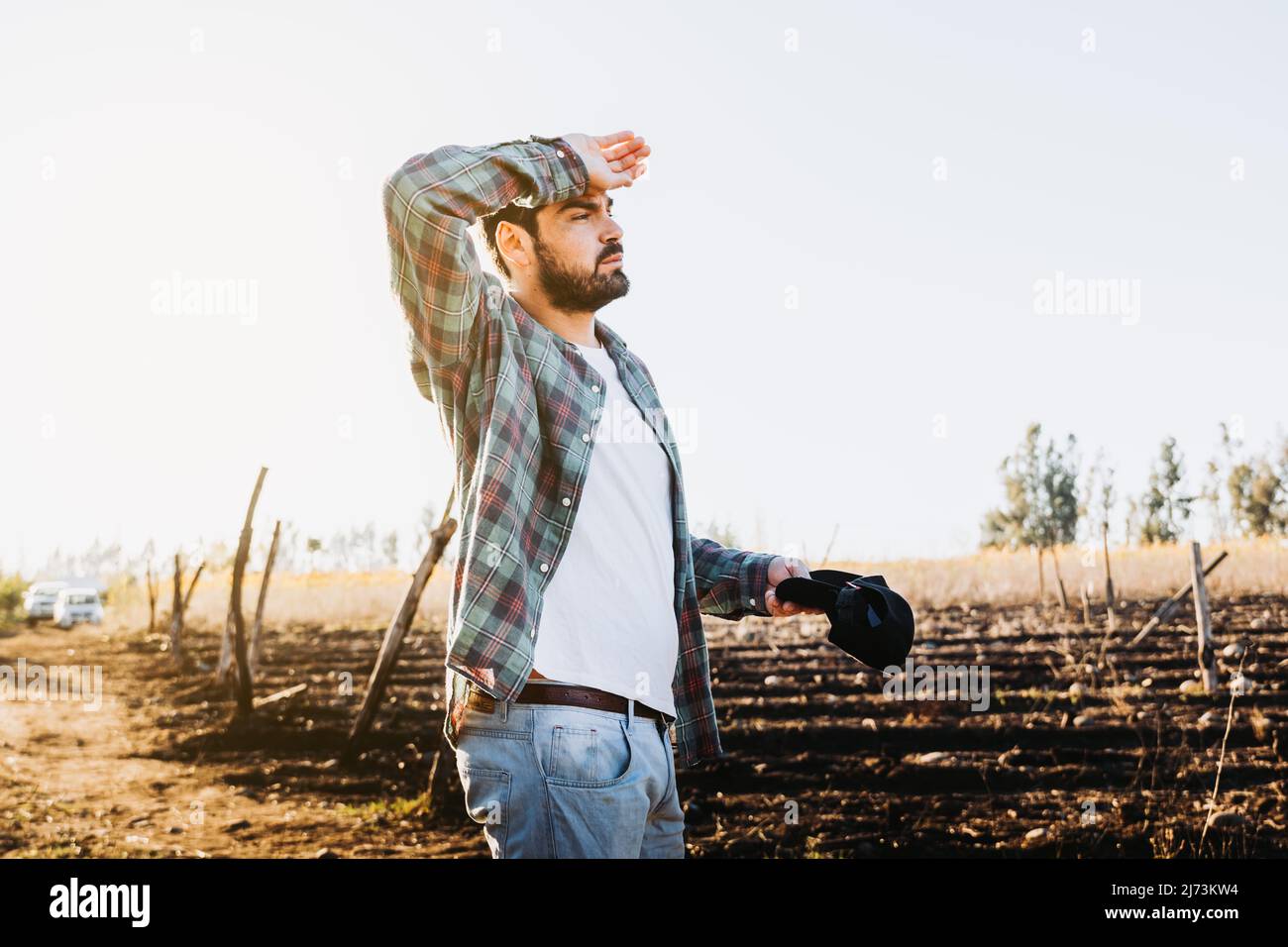 Giovane agricoltore lavoratore stanco dopo il lavoro, accanto al suo terreno agricolo e guardando le sue piantagioni. Foto Stock