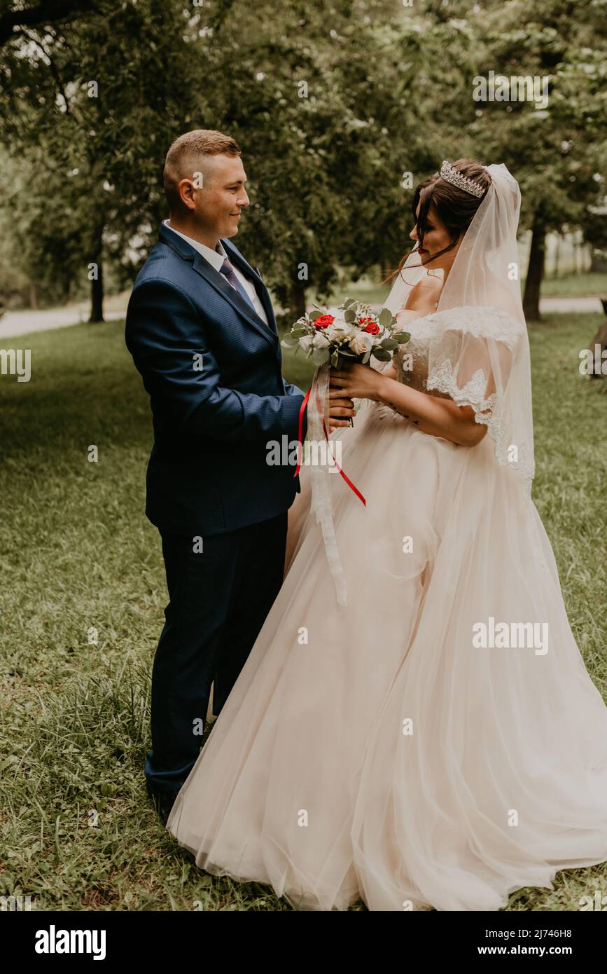 Il giovane uomo biondo caucasico europeo sposa in abito blu e donna dai capelli neri sposa in abito da sposa bianco con velo lungo e tiara sulla testa. Prima Foto Stock