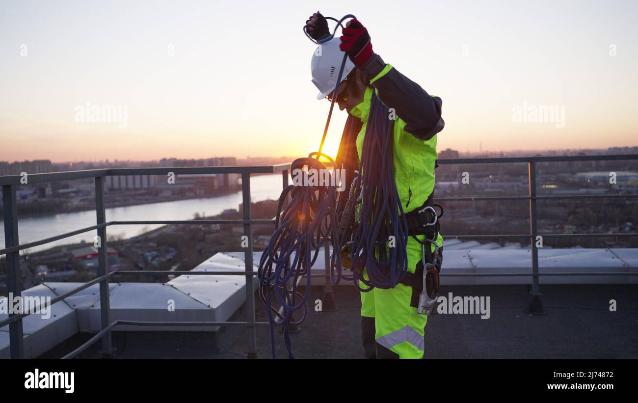 Arrampicatore nervoso cerca di distangere le funi di sicurezza blu e appende spiedini sulle spalle sul tetto dell'edificio. Il climber industriale si prepara al lavoro al tramonto Foto Stock