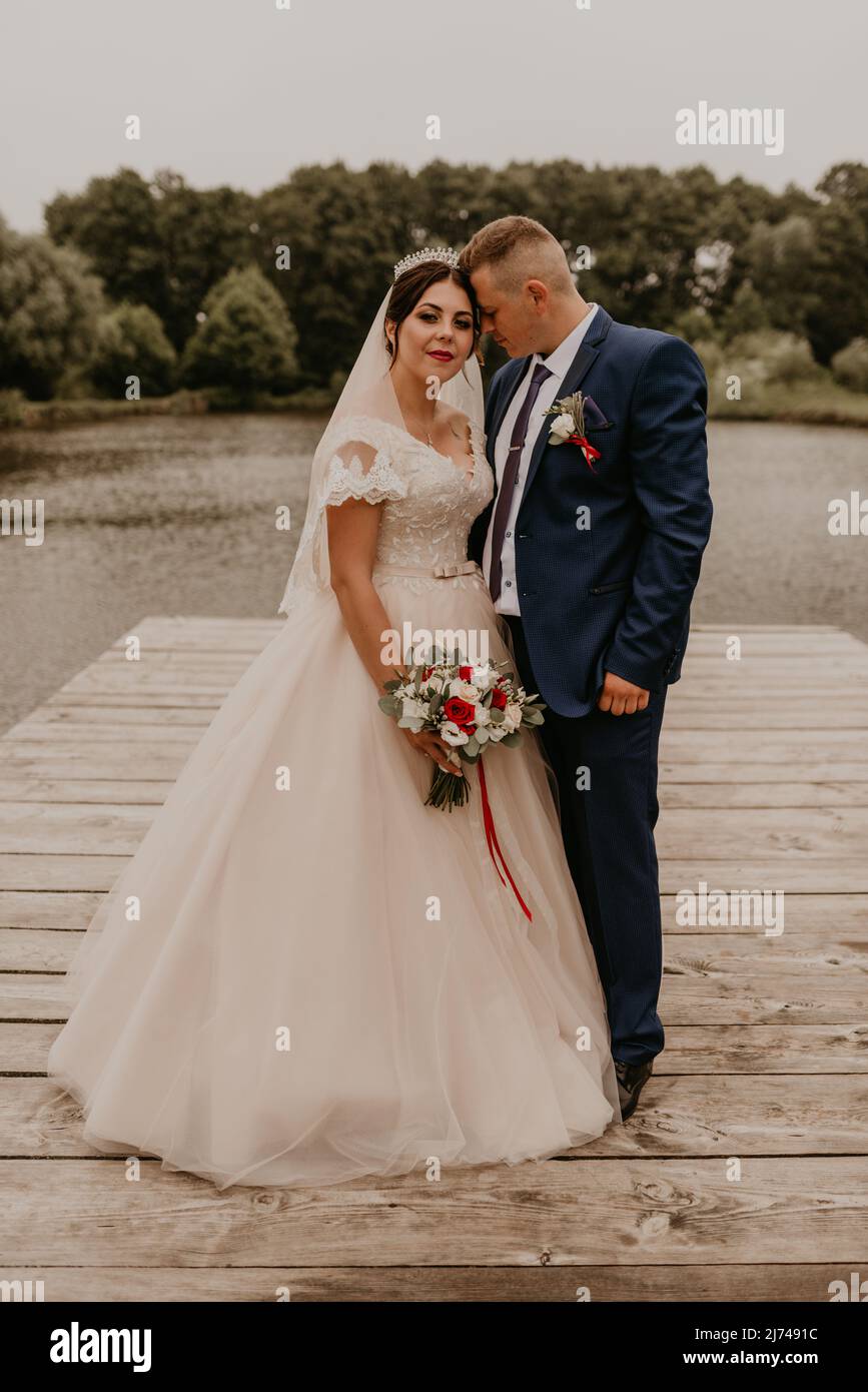 Il giovane uomo biondo caucasico europeo sposa in abito blu e donna dai capelli neri sposa in abito da sposa bianco con velo lungo e tiara sulla testa. Sposi novelli Foto Stock