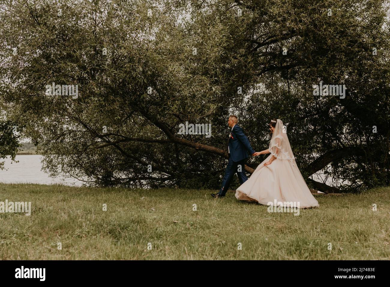 Il giovane uomo biondo caucasico europeo sposa in abito blu e donna dai capelli neri sposa in abito da sposa bianco a velo lungo e tiara sulla testa. Gli sposi camminano Foto Stock