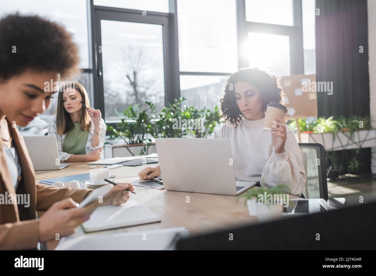 Donna d'affari musulmana che tiene il caffè per andare vicino ai colleghi e ai dispositivi interrazziali in ufficio Foto Stock