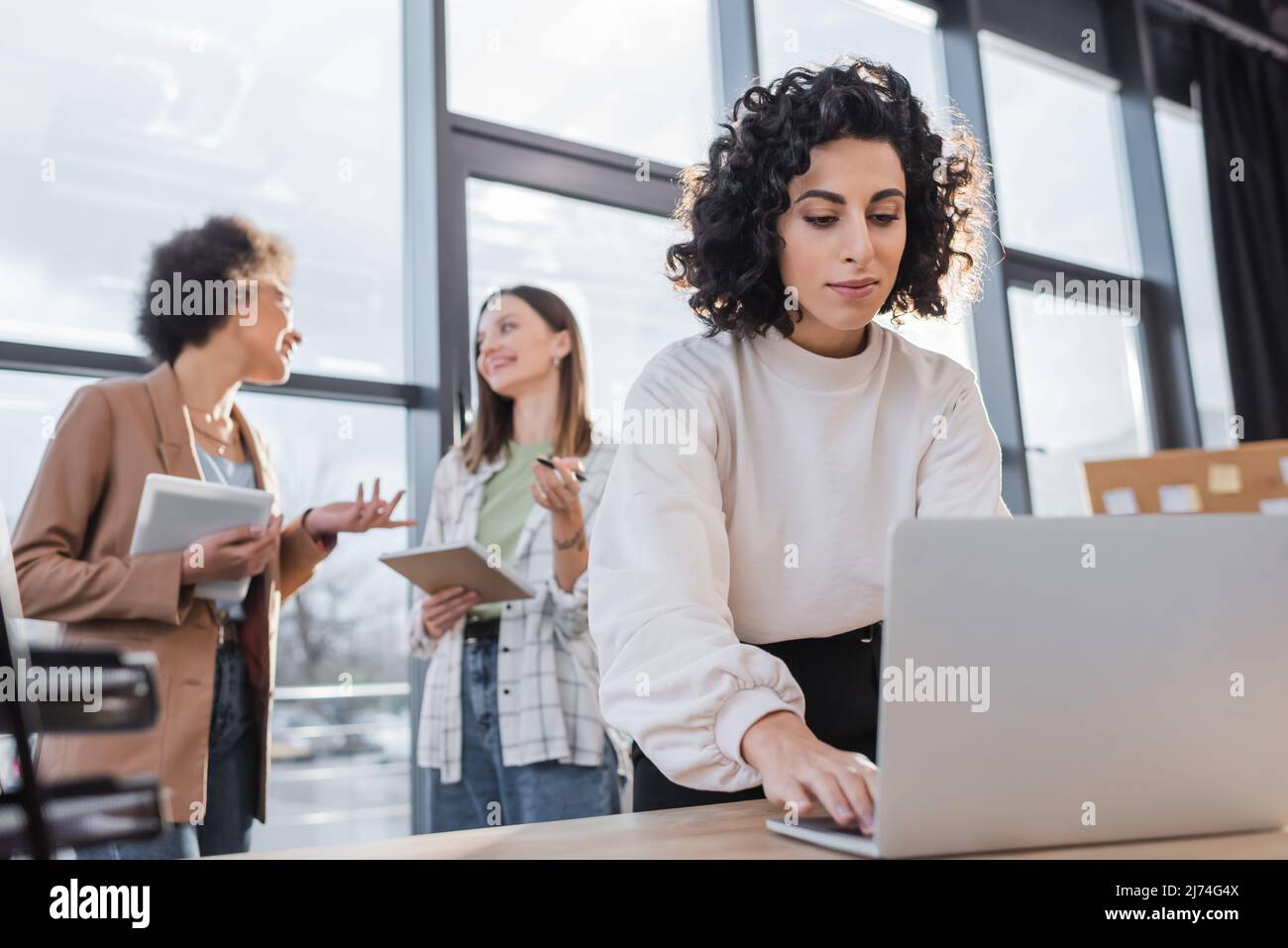 Donna d'affari musulmana che usa il laptop vicino ai colleghi multietnici offuscati che parlano in ufficio Foto Stock