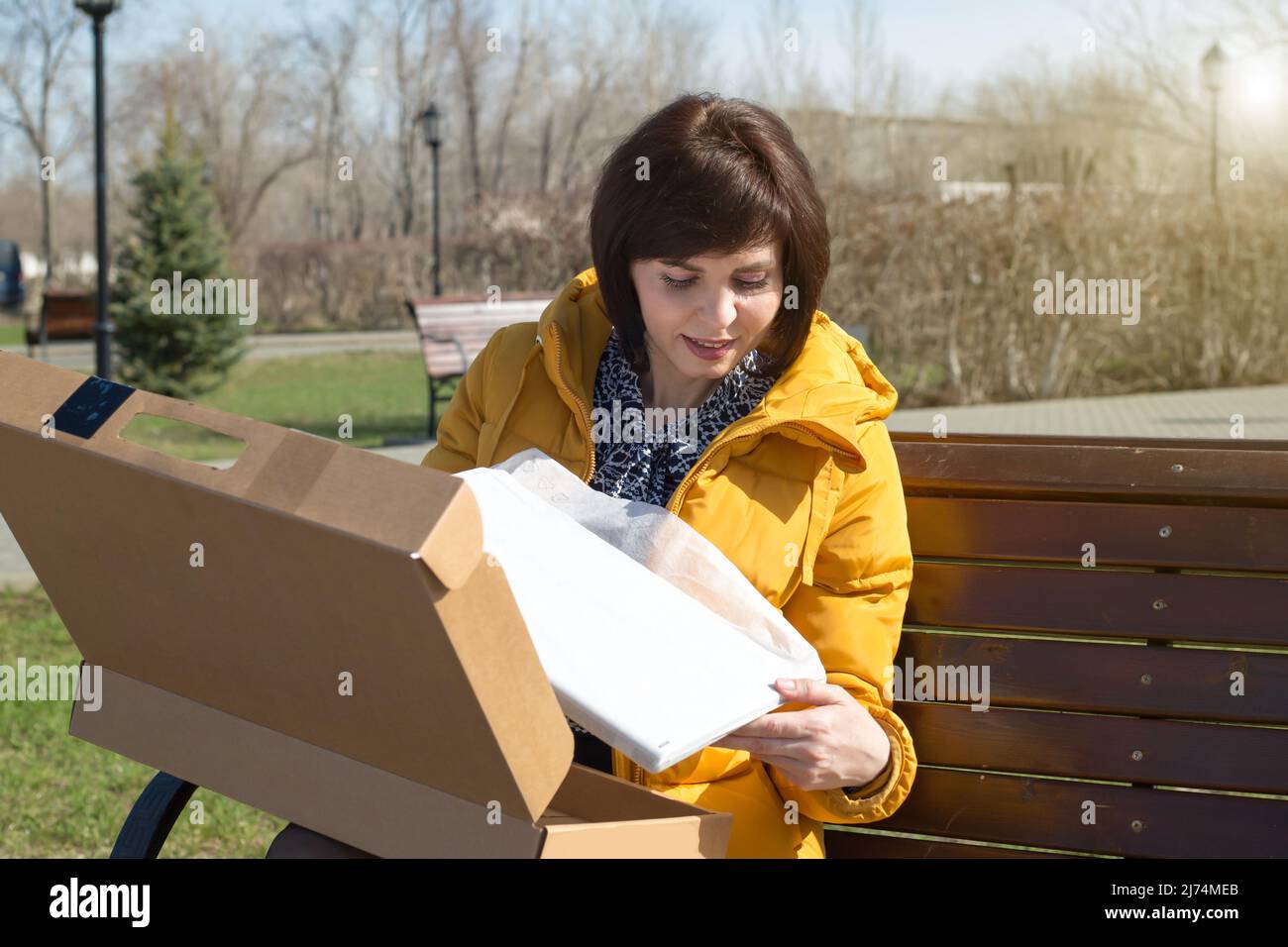 Una donna felice bruna in una giacca giallo brillante su una panca con un sorriso apre una scatola con un nuovo portatile. Foto Stock