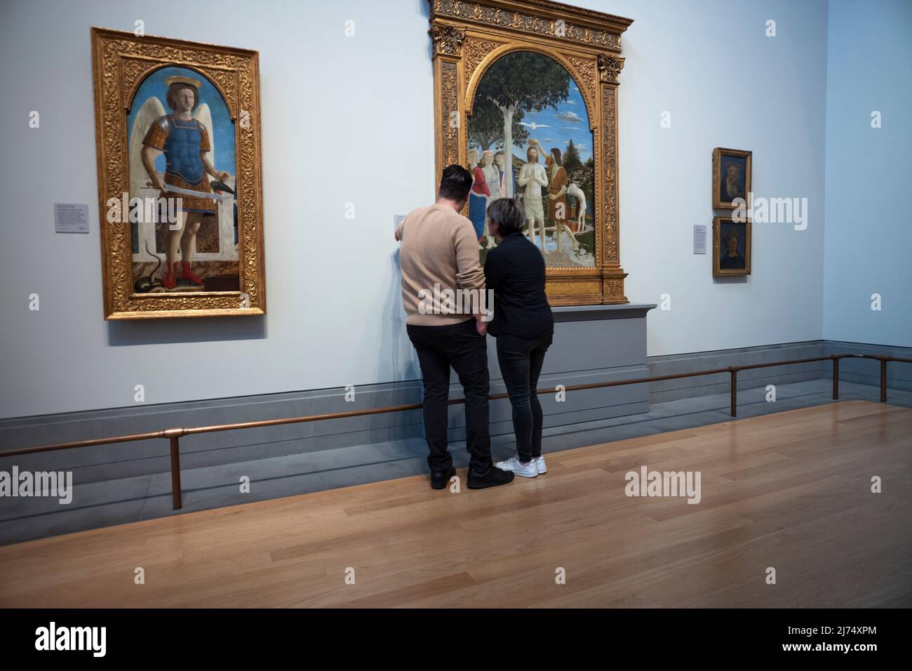 National Gallery London Inghilterra Aprile 2022 i visitatori tornano a Londra dopo i due anni di blocco di Covid che si sono visti qui alla National Gallery di Trafalgar Square nel centro di Londra. I visitatori ammirano: Il Battesimo di Cristo (centro) e San Michele (a sinistra) è un dipinto del maestro rinascimentale italiano Piero della Francesca. Dipinta in tempera d'uovo su due pannelli di legno di pioppo, la datazione è controversa – alcuni gli danno una data molto presto, forse 1439; altri molto più tardi, intorno al 1460. Si tiene presso la National Gallery di Londra. La composizione è centrata sulla figura dell'essere di Cristo Foto Stock