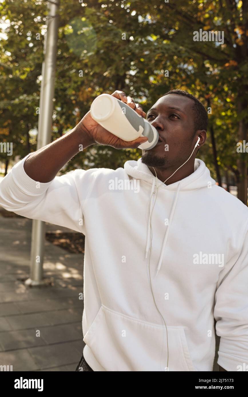 Giovane atletico uomo nero sete indossando la felpa bianca che beve l'acqua dalla bottiglia di sport dopo le lunghe distanze che corre all'aperto Foto Stock