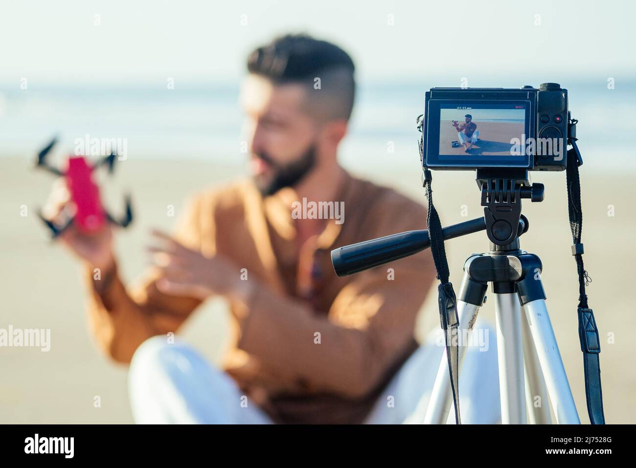 uomo arabo seduto sulla sabbia e parlare di treppiede video facendo revisione di quadrocopter in spiaggia. Concetto di creatore di contenuti lifestyle per i videografi di viaggio Foto Stock