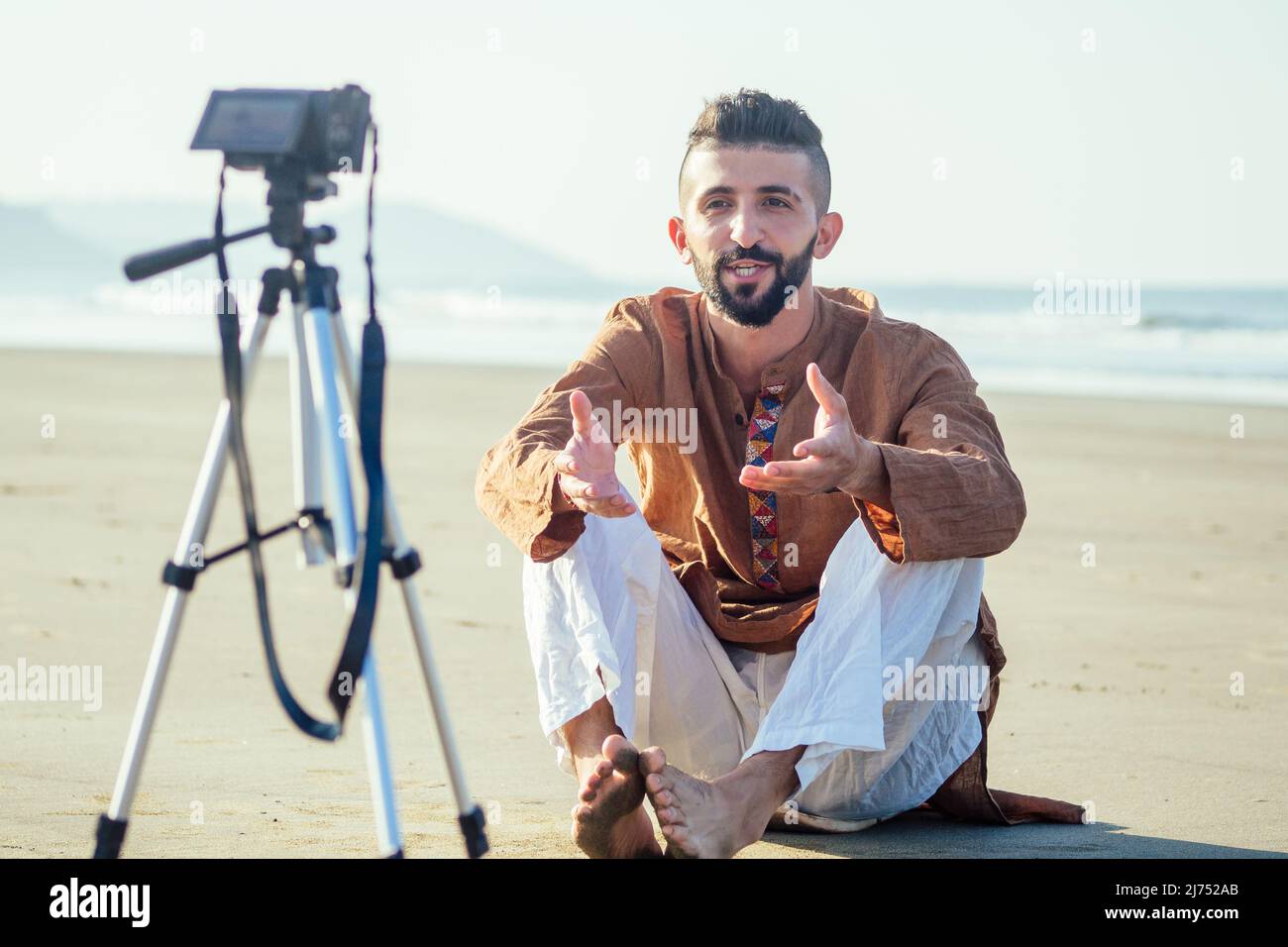 uomo arabo seduto sulla sabbia e parlare di tripode video fare in spiaggia. Concetto di creatore di contenuti lifestyle per i videografi di viaggio Foto Stock