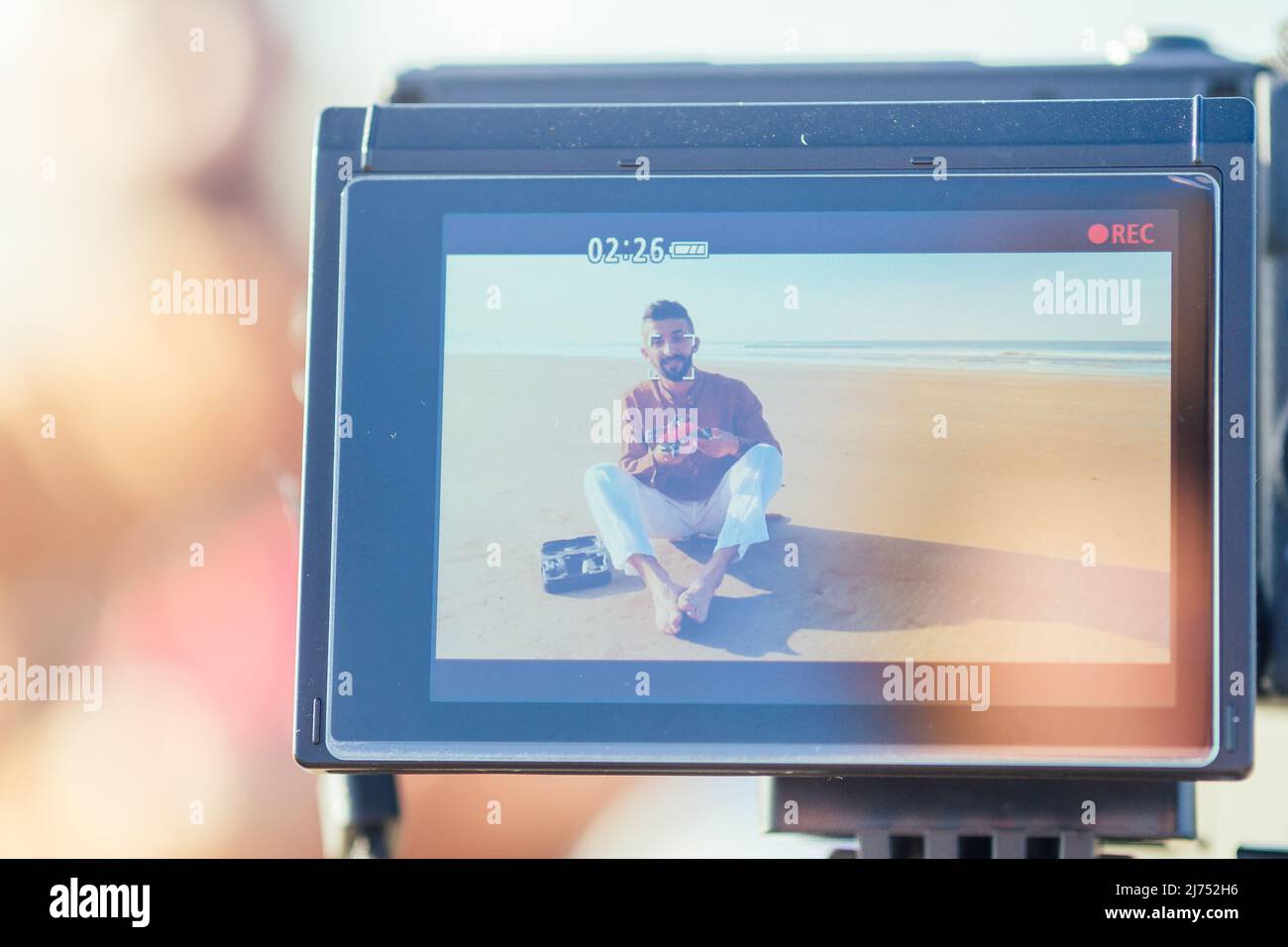 uomo arabo seduto sulla sabbia e parlare di tripode video fare in spiaggia. Concetto di creatore di contenuti lifestyle per i videografi di viaggio Foto Stock