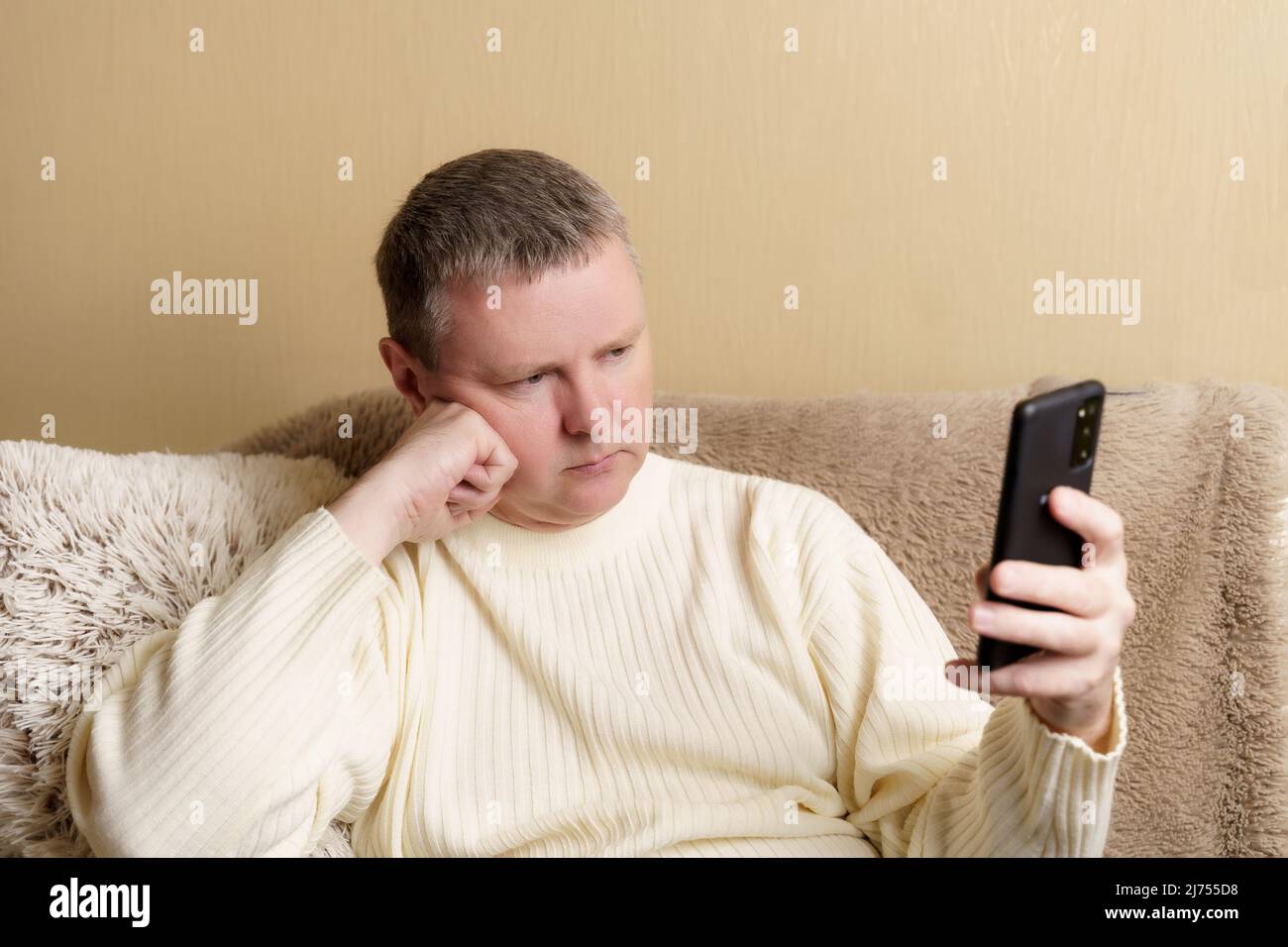 Un uomo guarda pensieroso in uno smartphone, poggiando la testa sulla mano. Europeo in un maglione bianco si siede su un divano con un telefono in mano. Foto Stock