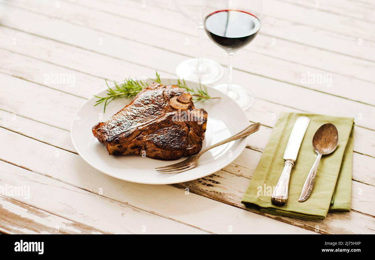 Bistecca intera su un piatto bianco con un rametto Rosemary con un bicchiere di vino rosso su un tavolo di legno bianco Foto Stock