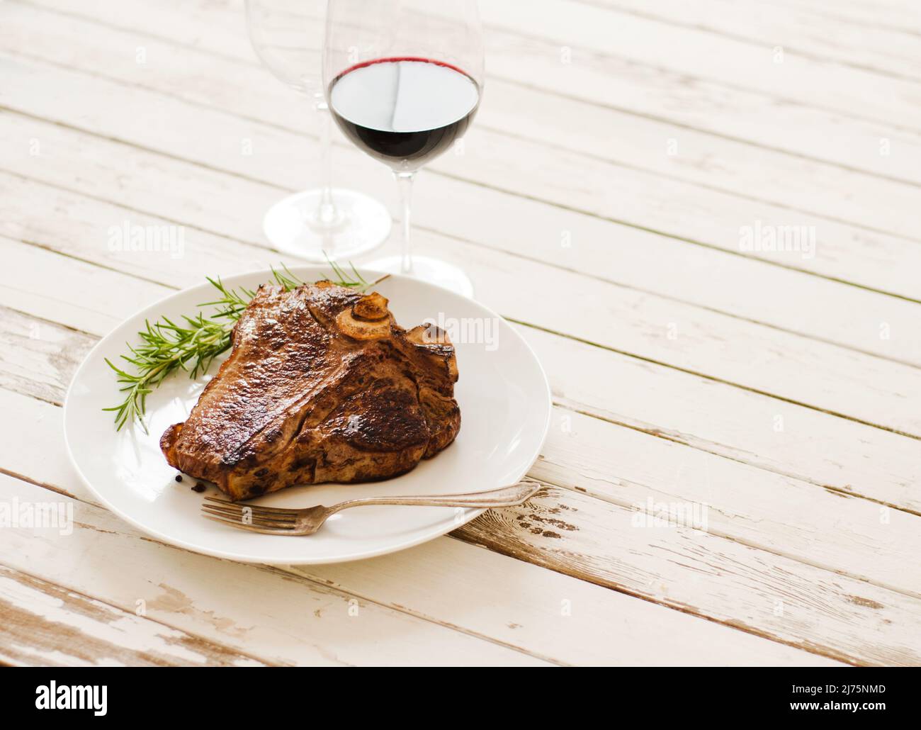 Bistecca intera su un piatto bianco con un rametto Rosemary con un bicchiere di vino rosso su un tavolo di legno bianco Foto Stock