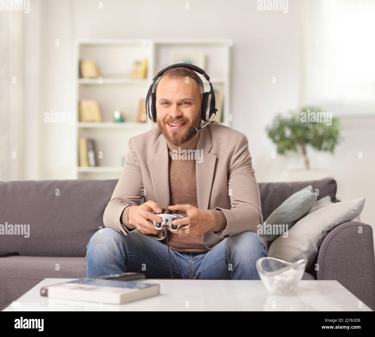 Giovane uomo con cuffie e un joystick seduto su un divano e giocare a videogiochi a casa Foto Stock