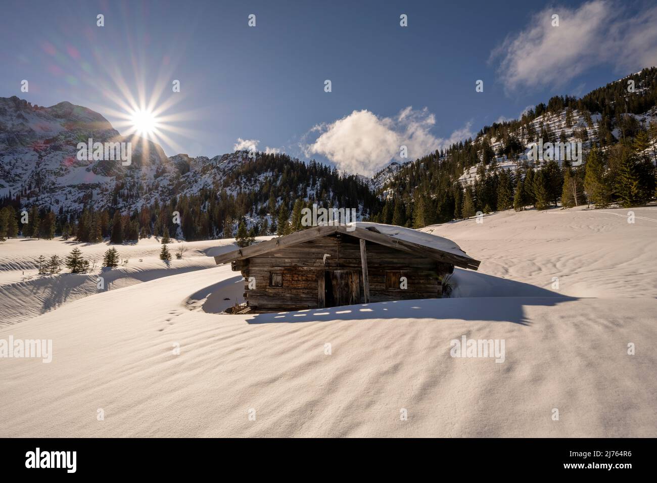 Una piccola capanna in legno nella Valle del Rodano, vicino a Hinterriss nel Karwendel, una parte delle Alpi in Austria in inverno con neve. Il sole è basso sulle montagne con le Karwendelspitze orientali, con un cielo prevalentemente blu e il tempo equo, la capanna getta la sua ombra verso lo spettatore, sullo sfondo il paesaggio nella neve. Foto Stock