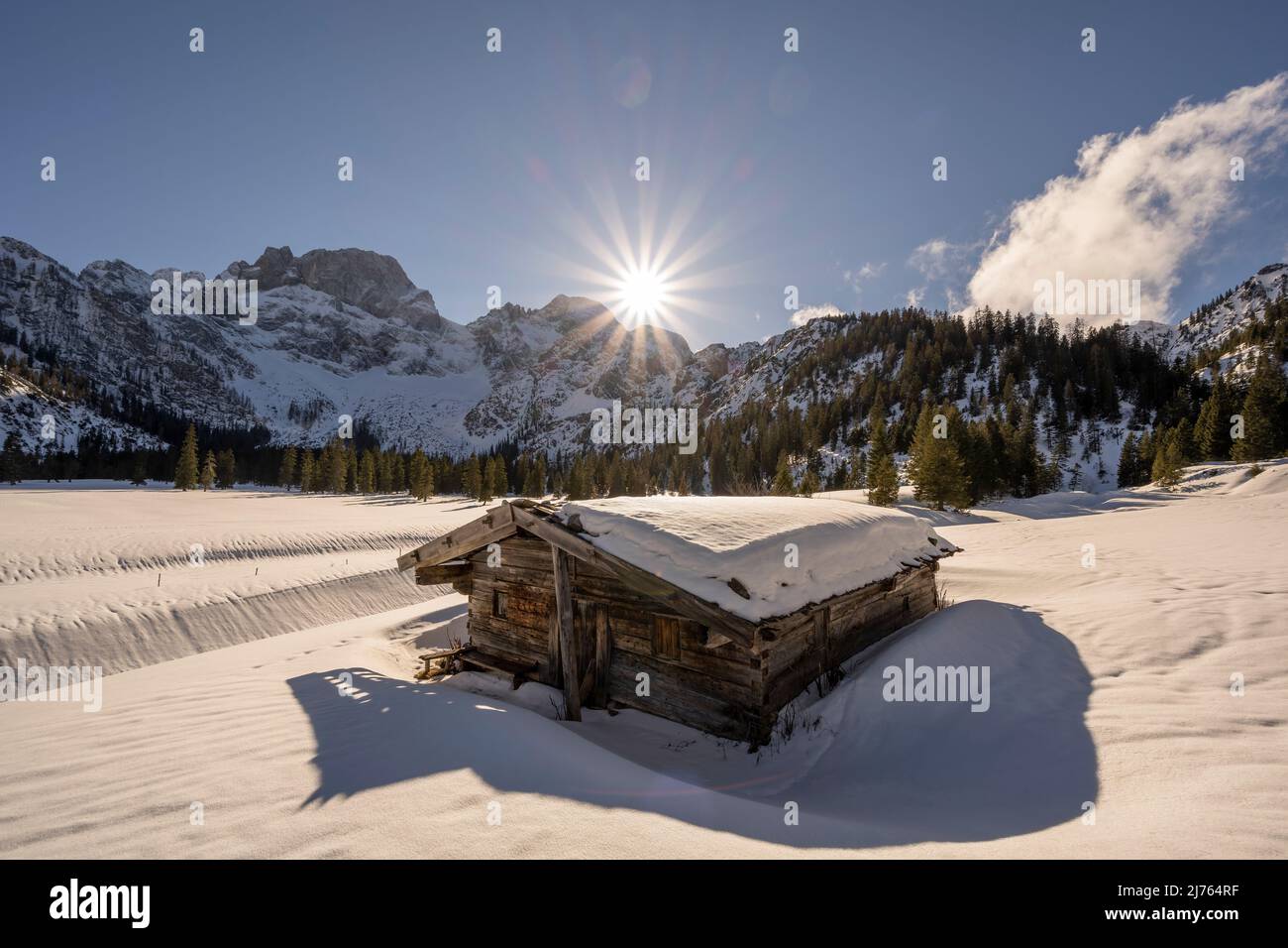 Una piccola capanna in legno nella Valle del Rodano, vicino a Hinterriss nel Karwendel, una parte delle Alpi in Austria in inverno con neve. Il sole è basso sulle montagne con le Karwendelspitze orientali, con un cielo prevalentemente blu e il tempo equo, la capanna getta la sua ombra verso lo spettatore, sullo sfondo il paesaggio nella neve. Foto Stock