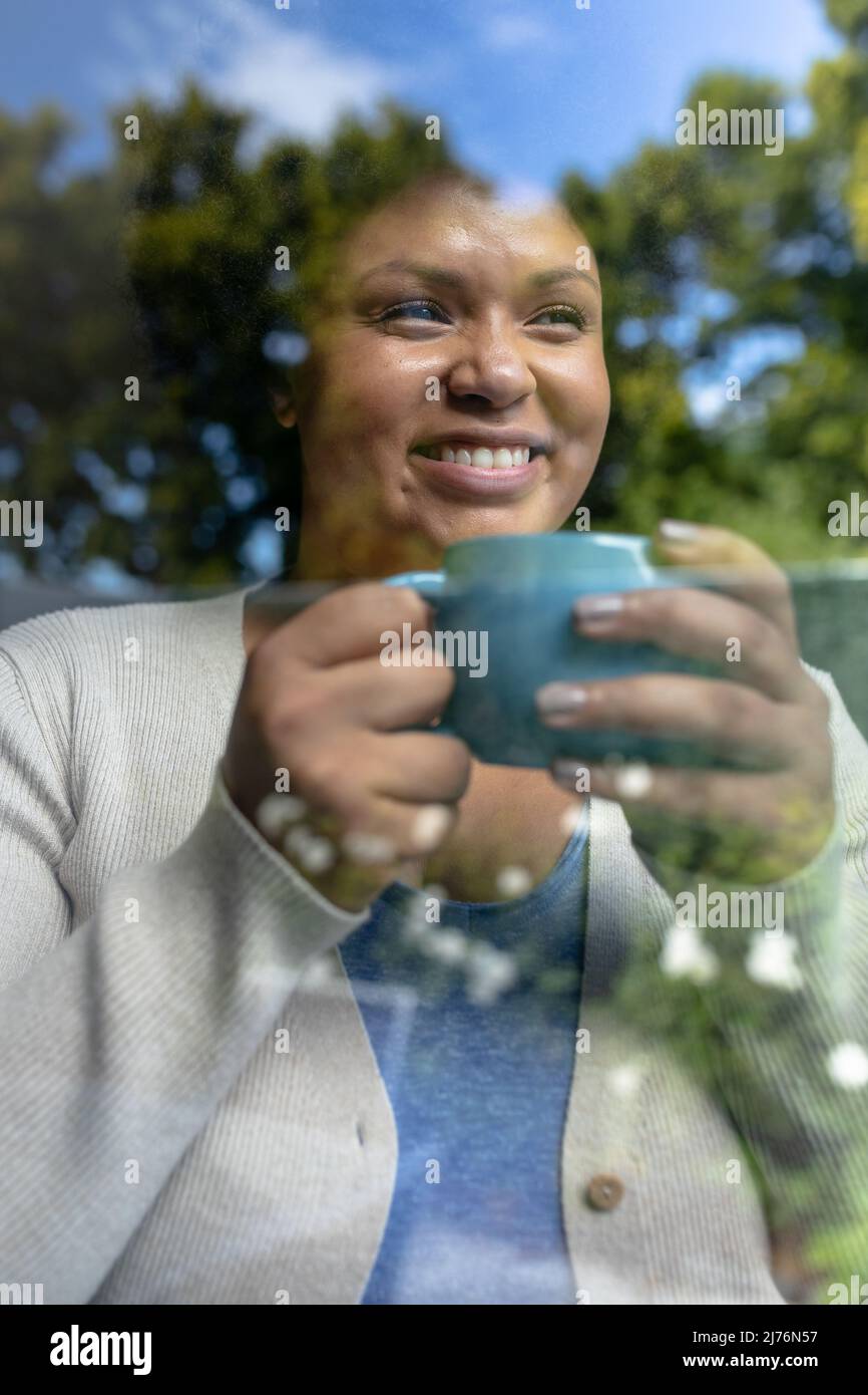 Donna adulta afroamericana sorridente con una tazza di caffè vista attraverso la finestra Foto Stock