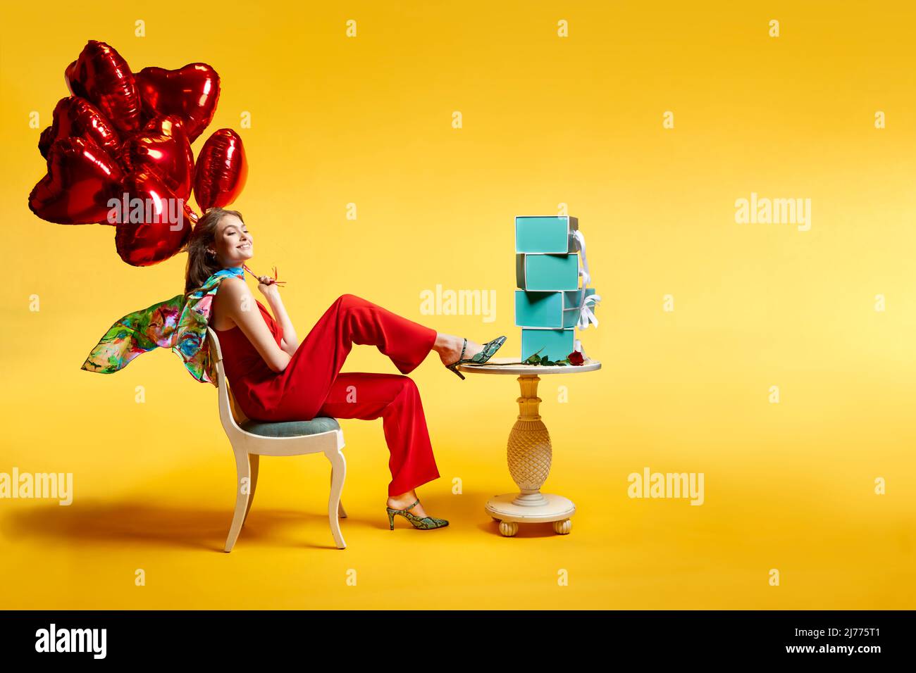 Una giovane donna felice in tiri eleganti tiene un bouqquet di palloncini a cuore con una pila di scatole di scarpe turchesi sul tavolo di fronte a lei. Foto Stock
