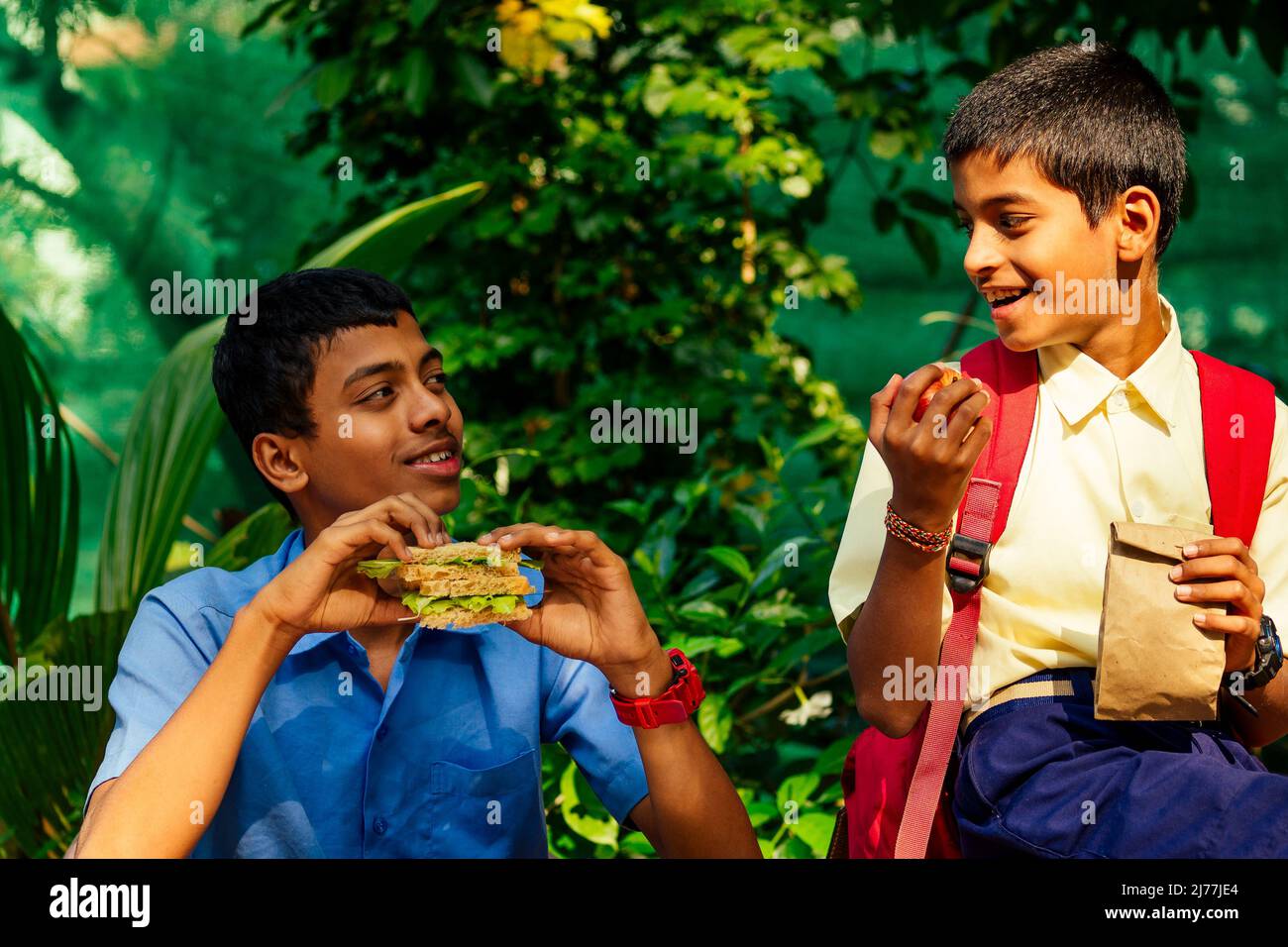 il ragazzo della scuola indiana che mangia sandwich e il suo amico con zaino si siede sul tavolo e mangia una mela nel parco Foto Stock