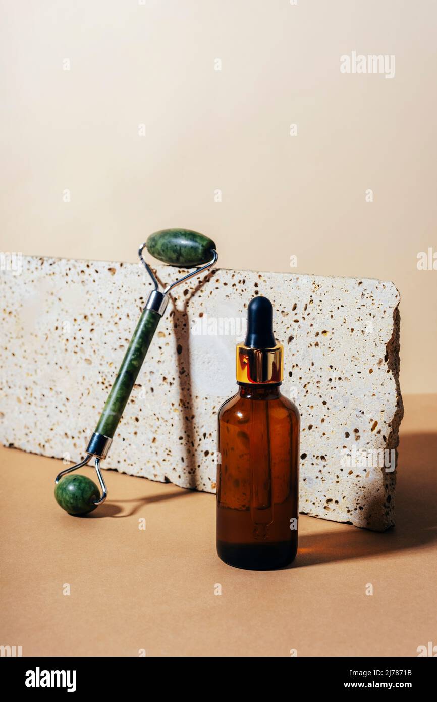 Bottiglia di siero e rullo facciale in giada su sfondo di pietra naturale con ombre dure. Primo piano. Foto Stock