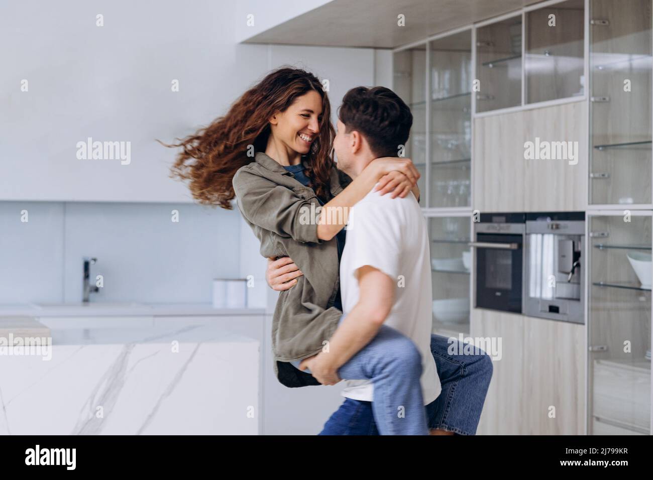 Giovane uomo brunetto cattura la tenuta in braccia e gira ricci moglie in camicia e jeans abbracciando strettamente e ridendo fortunatamente in cucina primo piano Foto Stock