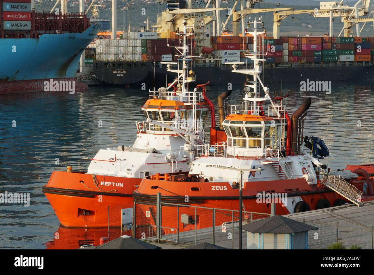 Due rimorchiatori con scafo arancione ormeggiati nel porto di Capodistria, Slovenia. Sullo sfondo sono presenti due contenitori in funzione cargo con gru a portale. Foto Stock