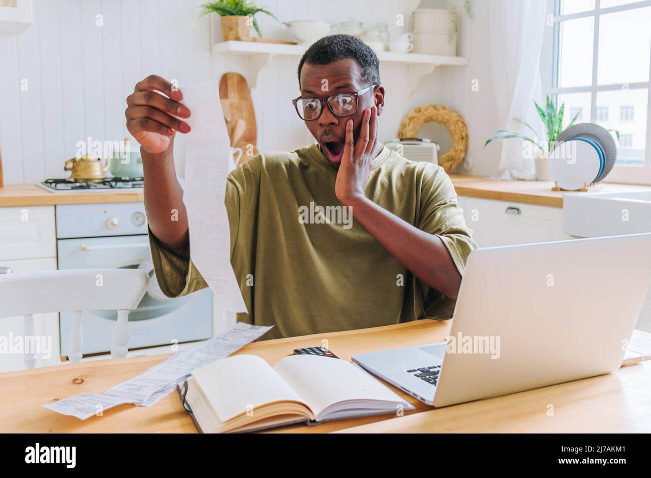 L'uomo afroamericano guarda gli assegni contando la quantità di denaro speso per lo shopping. Il ragazzo in occhiali si arrabbia seduto in cucina a casa vista ravvicinata Foto Stock