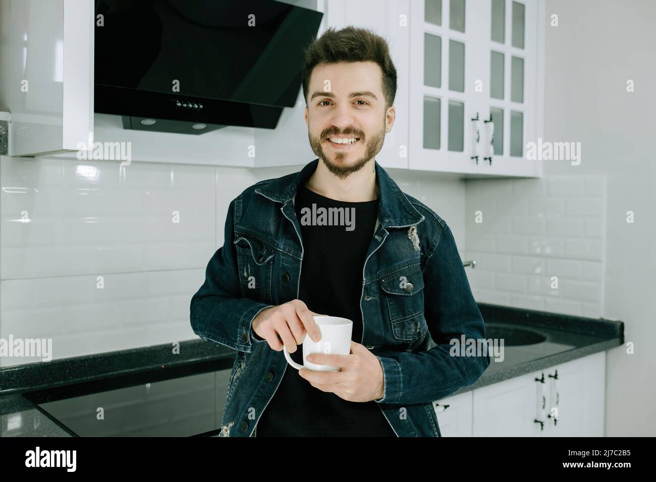 In posa, il giovane uomo sorride e tiene la tazza in cucina, il maschio adulto che indossa la giacca di denim che posa per la macchina fotografica Foto Stock