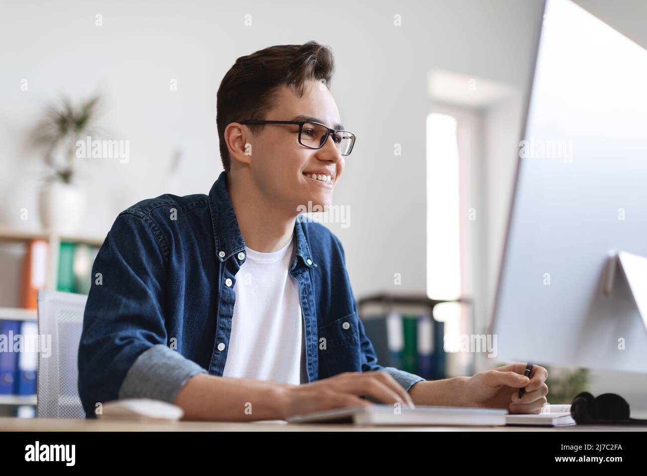 Giovane dipendente di sesso maschile che prende appunti mentre lavora sul computer alla scrivania Foto Stock
