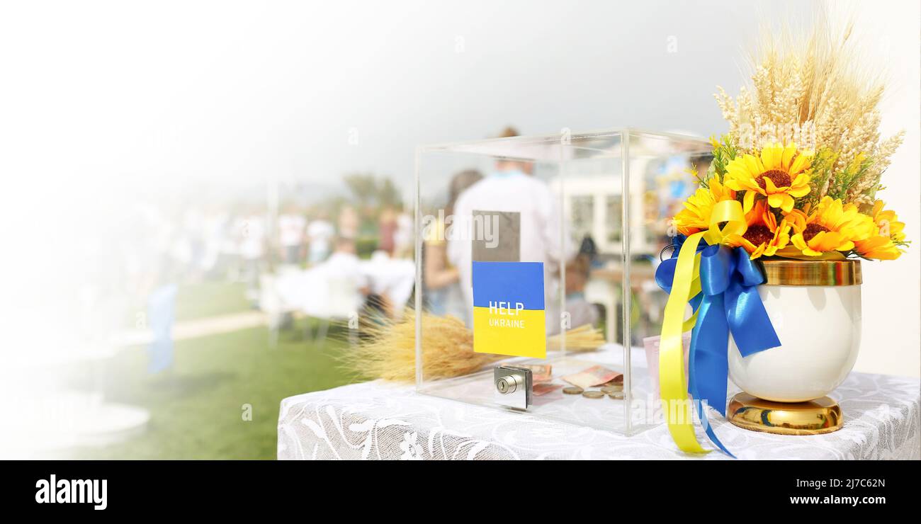 Beneficenza Ucraina raccolta fondi per donazioni in denaro in una scatola acrilica, vaso con fiori gialli e piante secche, nastro blu e giallo. Foto Stock