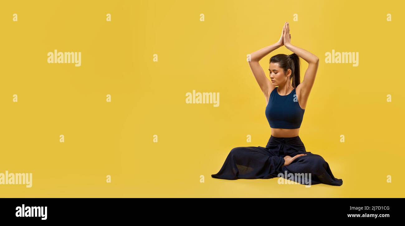 Panorama di bruna ragazza che pratica yoga posa in studio. Giovane donna snella seduta sul pavimento, tenendo le mani di preghiera sopra la testa, guardando verso il basso. Concetto di nuova età e yoga fare, Foto Stock