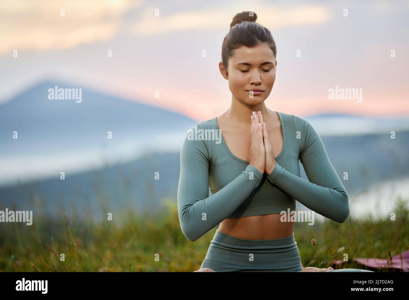 Vista frontale della donna brunetta che pratica la posizione del loto con gli occhi chiusi che mette le palme insieme sull'erba in montagne dell'alba. Raccolto di donna asiatica meditando sulla natura. Concetto di stile di vita sano. Foto Stock