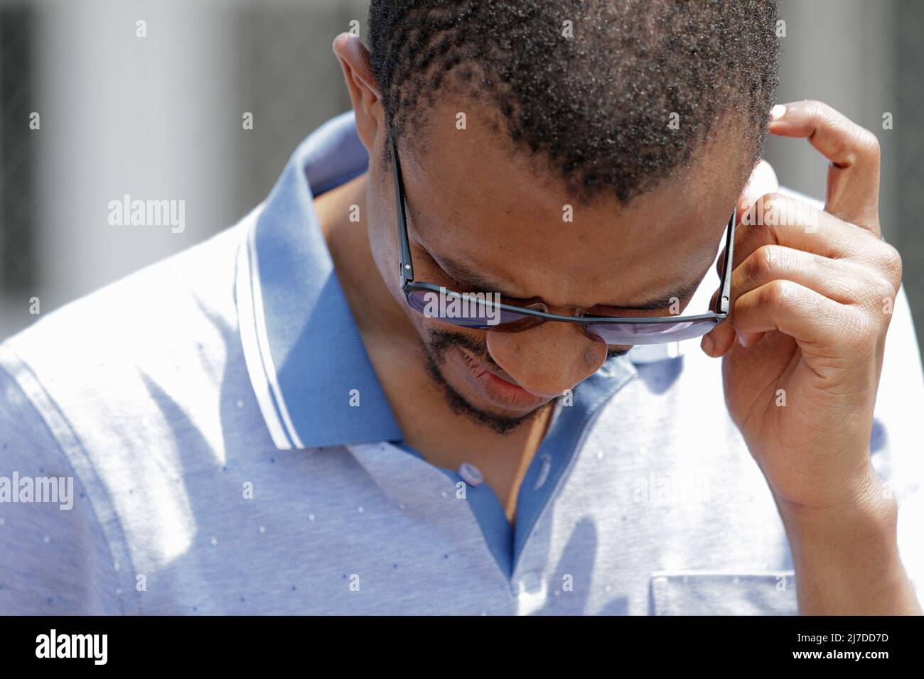 Uomo in profonda concentrazione, guardando in basso, graffiando la testa, indossando occhiali da sole e una camicia da golf blu. Foto Stock