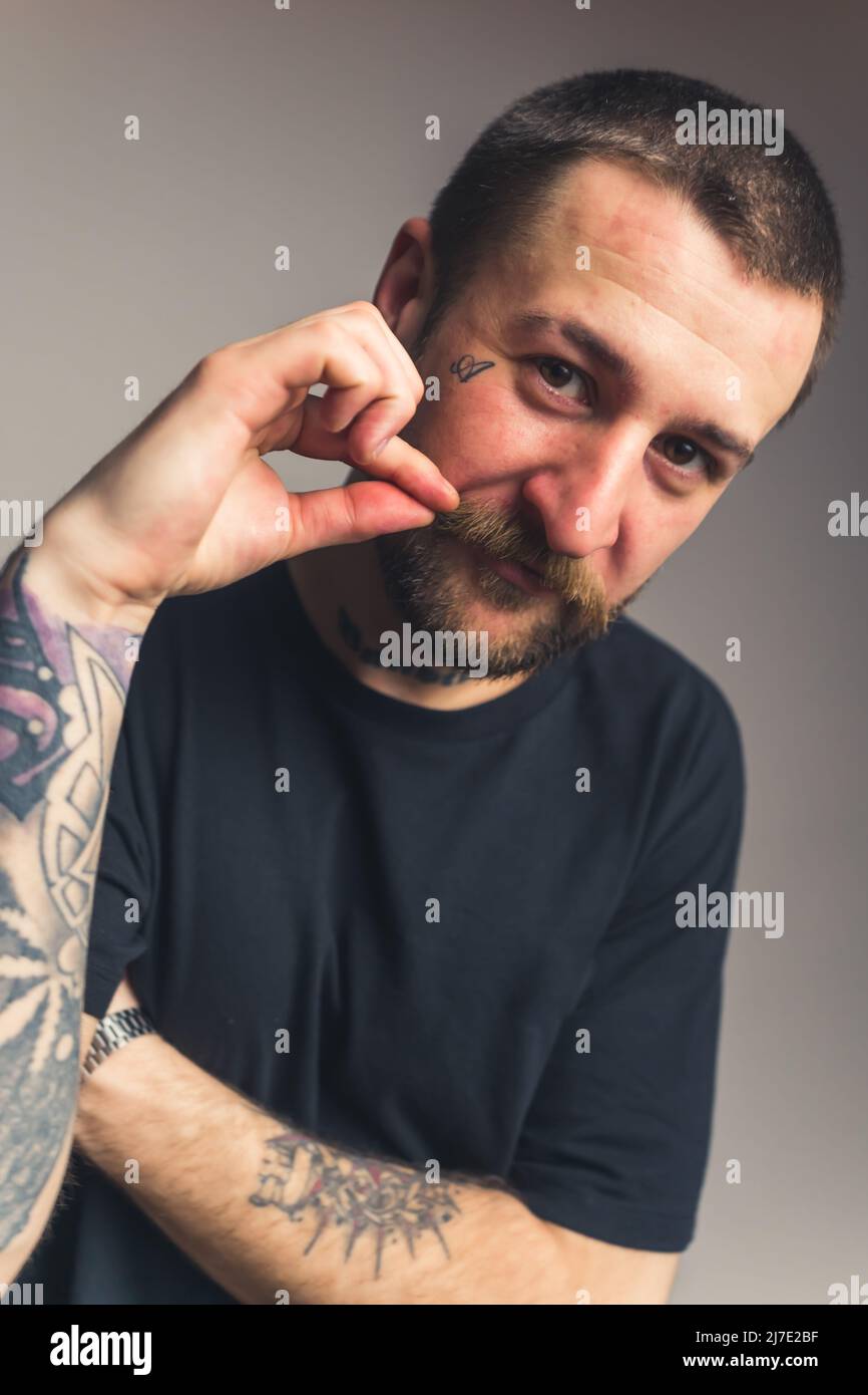 Ritratto verticale di un uomo tatuato di mezza età che tocca i baffi marroni e guarda la fotocamera su sfondo grigio. Foto di alta qualità Foto Stock