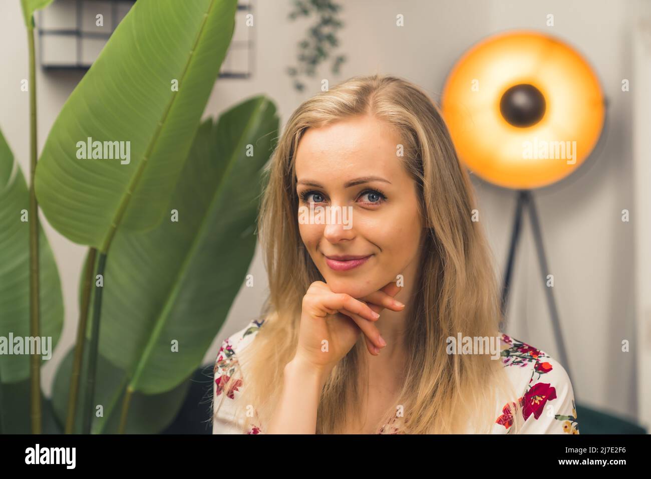 Closeup medio di donna caucasica bionda in blusa floreale, che sostiene il mento con mano salotto sfondo spazio copia . Foto di alta qualità Foto Stock