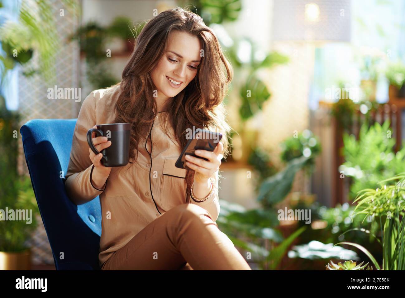 Casalinga sorridente di mezza età con capelli ondulati lunghi con tazza di cioccolata calda in pantaloni beige e blusa invio di SMS con smartphone Whi Foto Stock