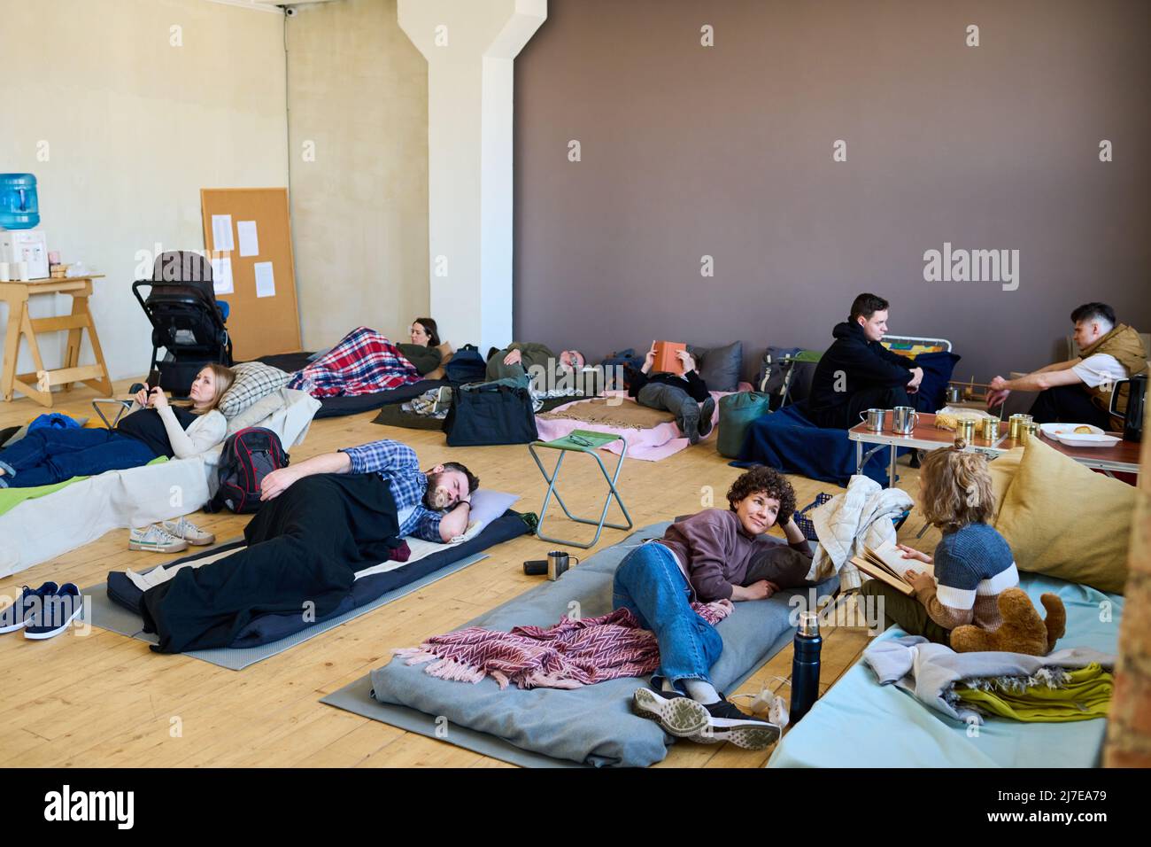 Diversi rifugiati che riposano sui loro posti letto su pavimento di legno in una camera spaziosa, mentre la giovane donna che parla con suo figlio con libro Foto Stock