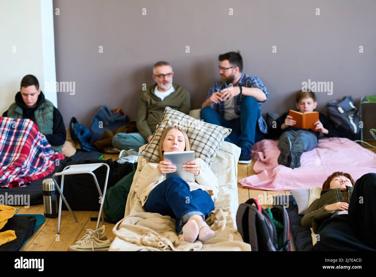 Giovane donna bionda che usa la tavoletta contro un gruppo di uomini che parla sul letto mentre riposa sul posto letto preparato per i migranti Foto Stock