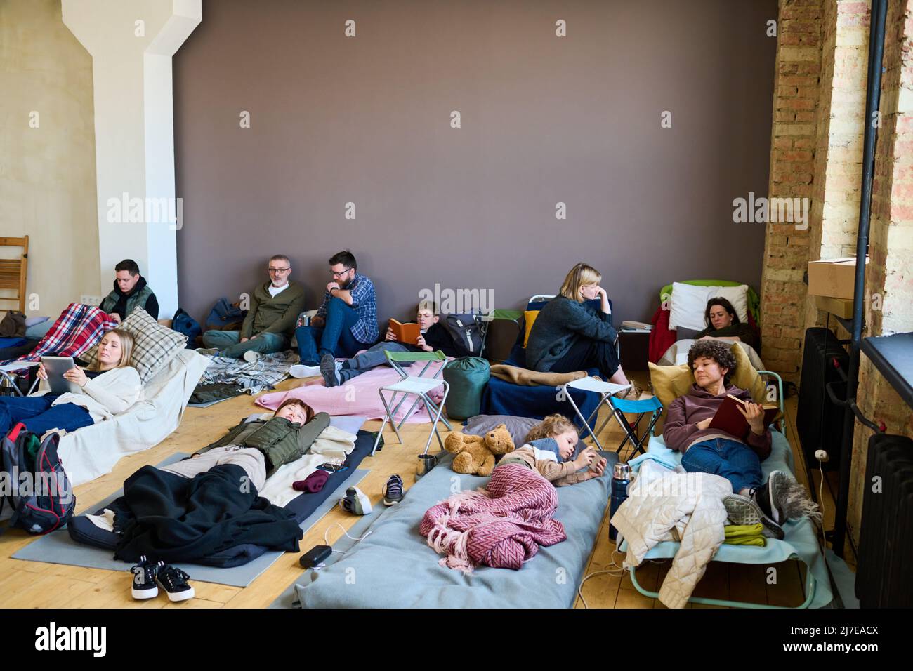 Gruppo di rifugiati che utilizzano gadget mobili, che parlano e dormono mentre si riposano su cuccette e materassi in una camera spaziosa Foto Stock